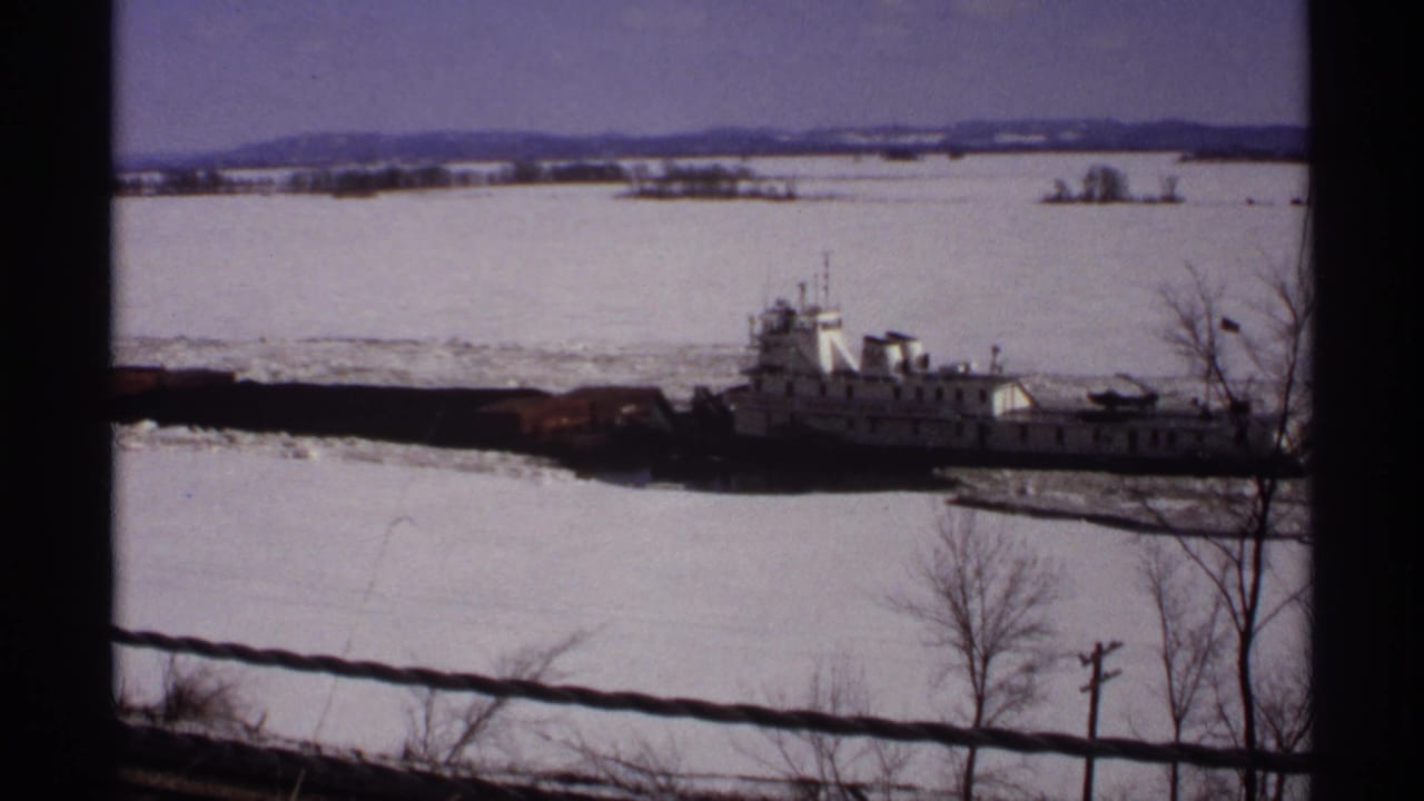 Icebreaker tug continues across a frozen river near bare trees.