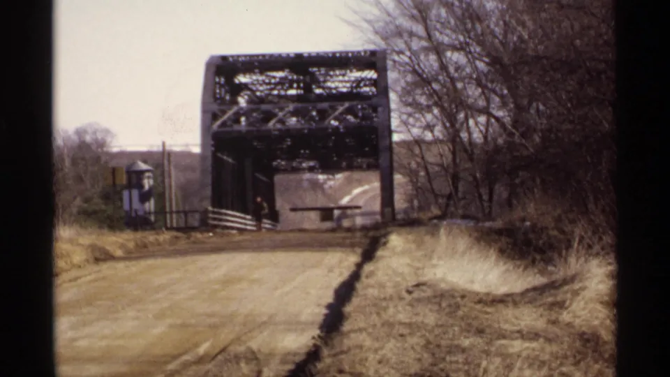 1971 Wisconsin 8mm frame of a rural steel truss bridge over a dirt road, with one distant person near the bridge entrance.