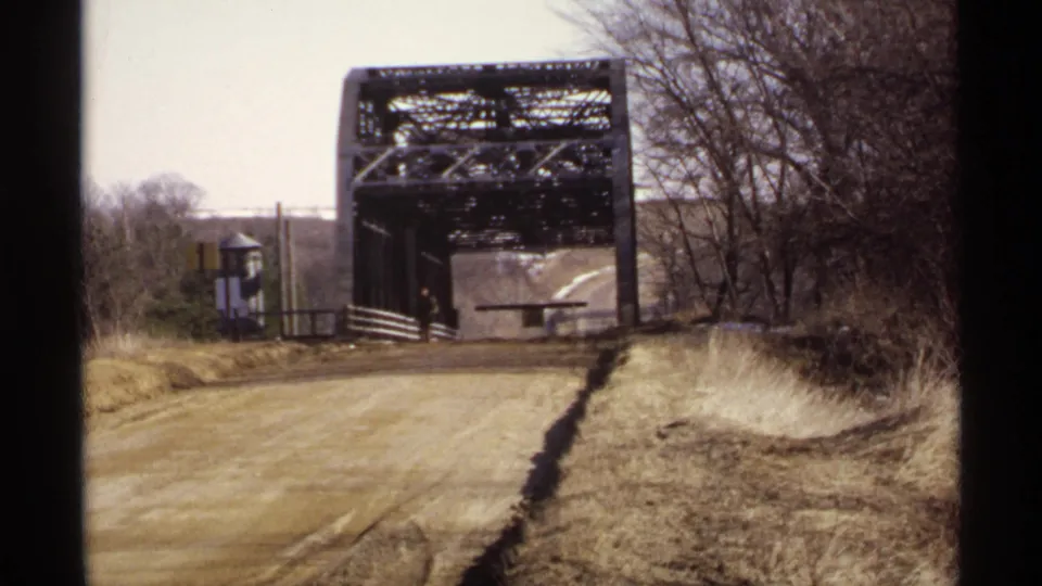 1971 Wisconsin home-movie frame showing a weathered steel truss bridge and dirt road, with bare trees and roadside grass nearby.