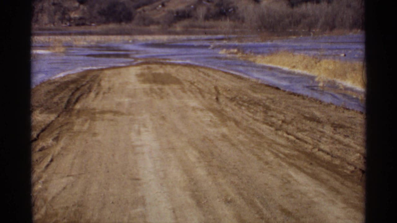 1971 Wisconsin rural road scene with a dirt surface, roadside water, dry grasses, and leafless trees in a quiet landscape.