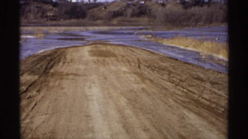 Wisconsin 1971 8mm frame of a dirt road running along a wet roadside area with dry reeds, brush, and bare trees.
