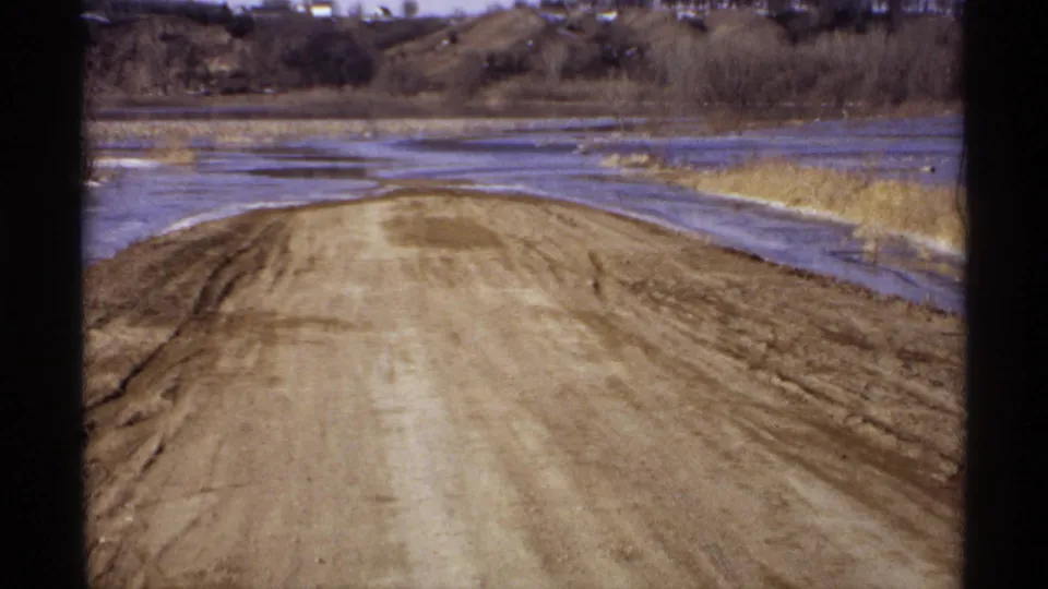 Archival 1971 Wisconsin home-movie frame showing a dirt road bordered by shallow water, dry reeds, and leafless trees.