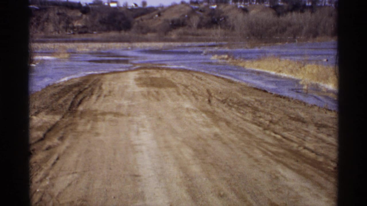 1971 Wisconsin 8mm frame showing a muddy dirt road along a low lakeshore with exposed reeds, dry grass, bare trees, and distant hills.