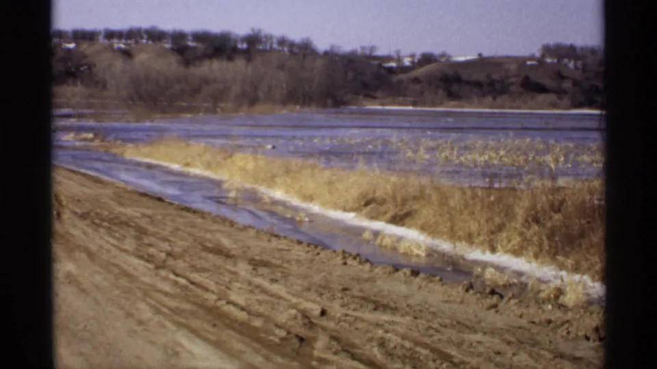 1971 Wisconsin 8mm frame with a curving muddy road beside shallow lake water, yellowed grass, reeds, bare trees, and distant terrain.