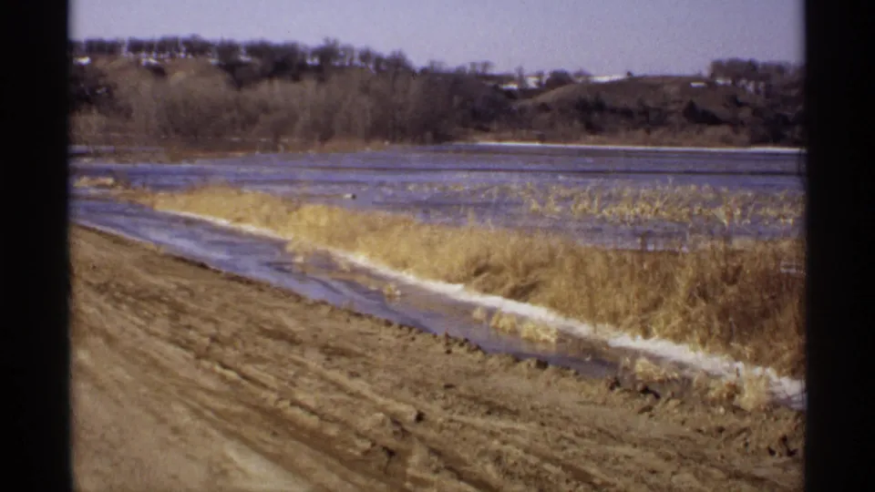 1971 Wisconsin 8mm view of a muddy road shoulder and dry shoreline grass beside low water, with reeds, bare trees, and distant hills.