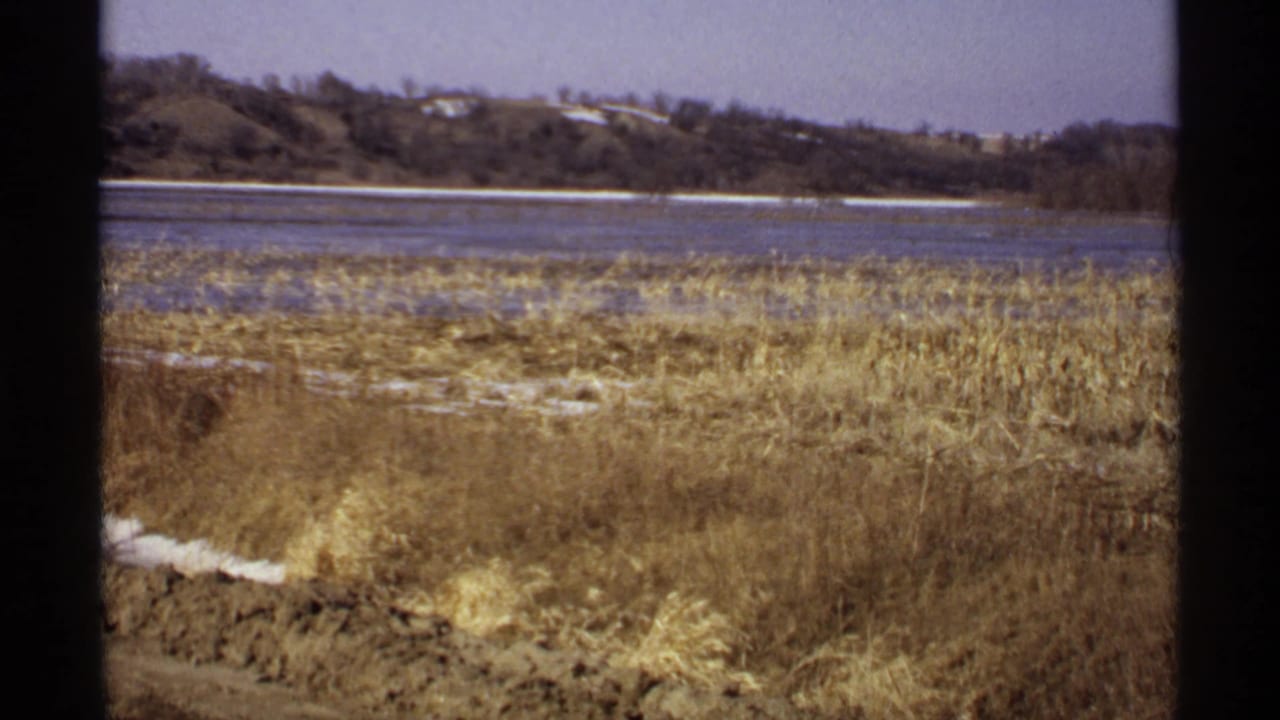 1971 Wisconsin 8mm frame showing dormant lakeside grass, small snow patches, calm water, and a distant wooded shore.