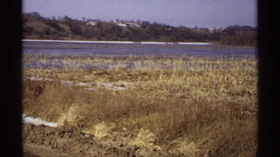 1971 Wisconsin 8mm view across dry reeds and patchy snow toward a calm lake and wooded hillside.