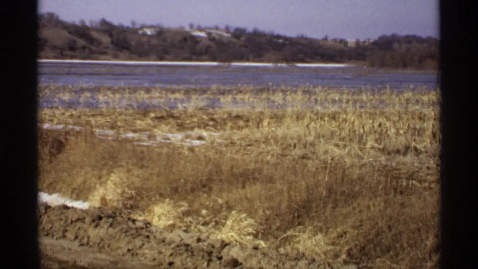 1971 Wisconsin home-movie frame with dry lakeside grass, lingering snow, calm water, and a tree-lined shore.