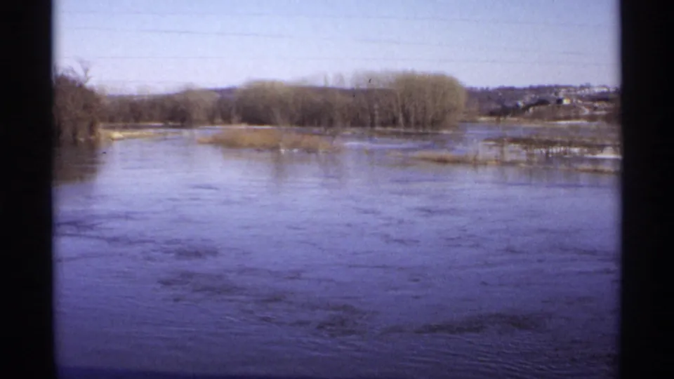 A 1971 Wisconsin home-movie view looks across a broad river toward bare trees, low banks, and faint distant buildings beyond the water.