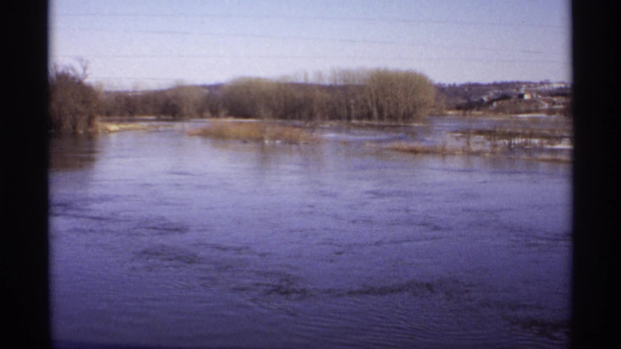 1971 Wisconsin 8mm frame showing broad floodwater, slow currents, bare riverside trees, and a distant rural shoreline.