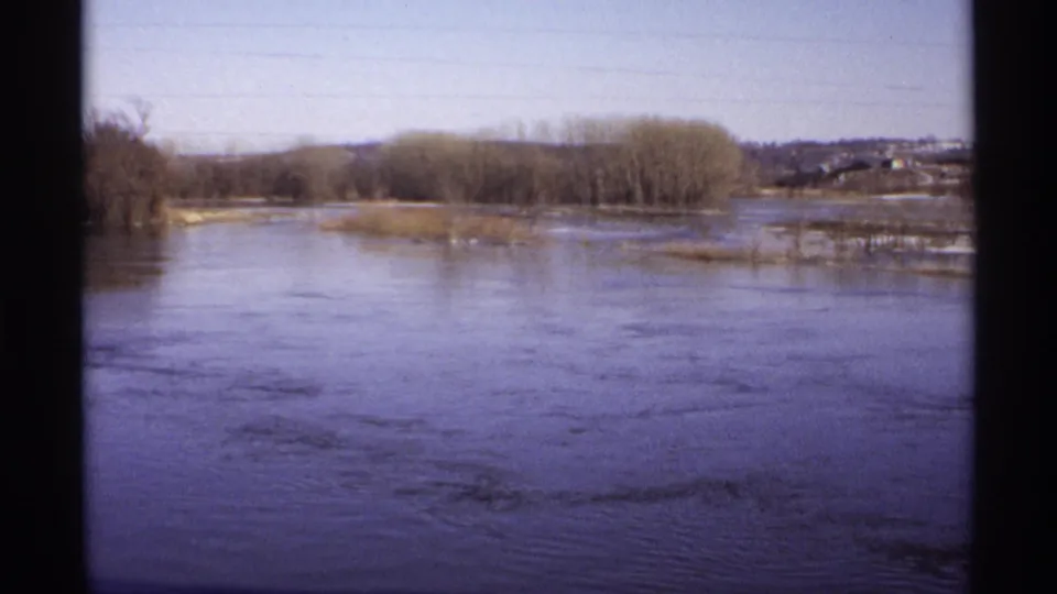 1971 Wisconsin 8mm frame showing broad floodwater, slow currents, bare riverside trees, and a distant rural shoreline.