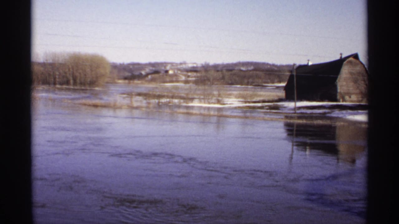 1971 Wisconsin 8mm frame showing floodwater over rural fields, bare trees, utility lines, and a dark barn near the water.