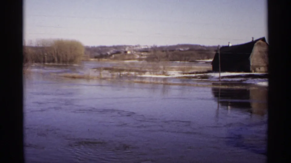 1971 Wisconsin 8mm frame showing submerged rural ground, a barn near the right side, bare trees, and distant hills under a pale sky.