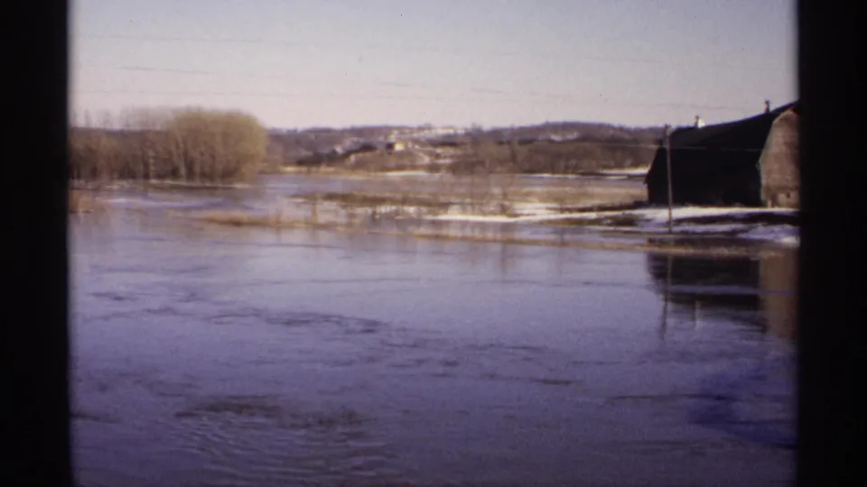 1971 Wisconsin 8mm frame showing slow flood currents, a partially surrounded barn, bare trees, and utility wires across rural land.