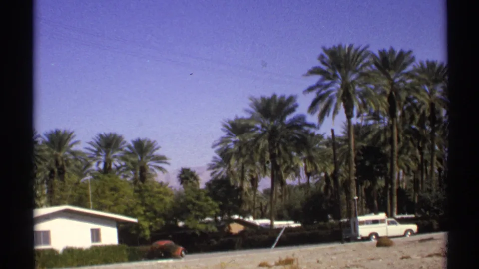 1972 Arizona 8mm frame with a low house, tall palms, a red car, and a white station wagon beside a gravel foreground.