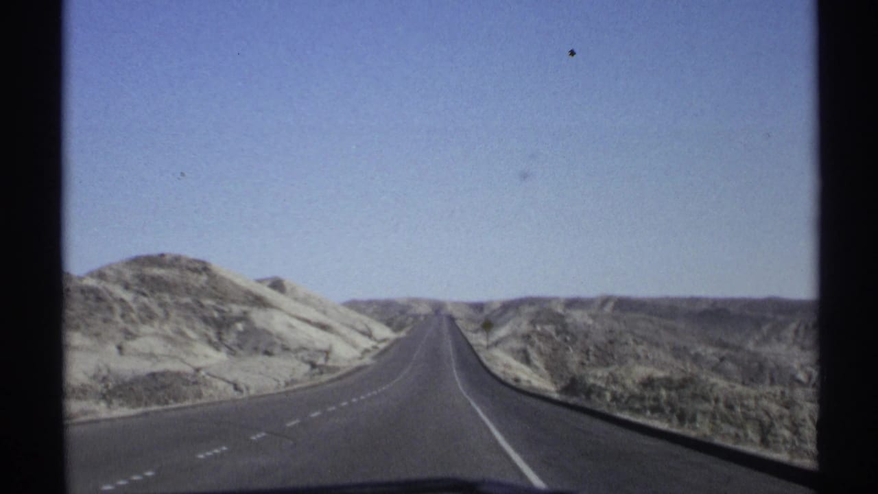 1972 Arizona 8mm travel view from a vehicle on a straight road through pale desert hills.