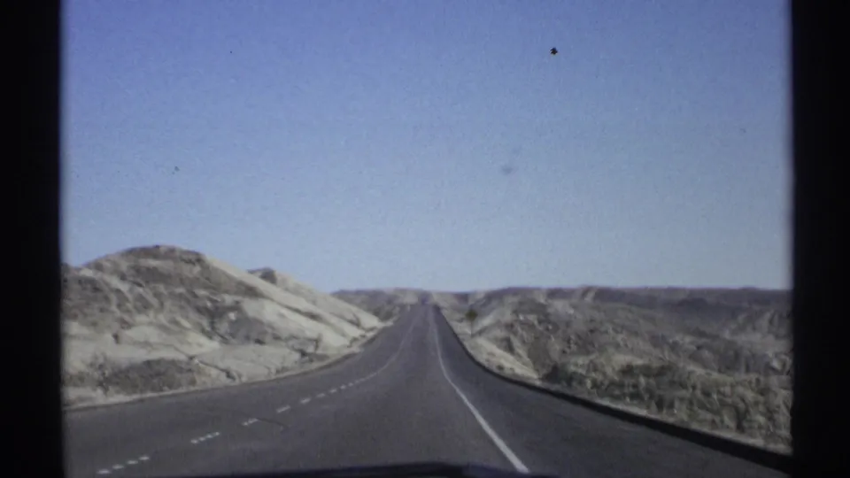 1972 Arizona 8mm travel view from a vehicle on a straight road through pale desert hills.