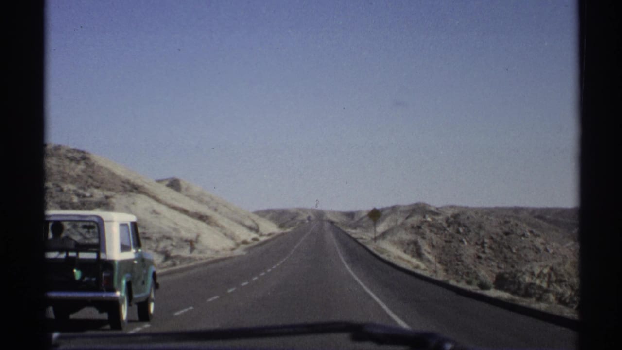 Green and white station wagon driving on a two-lane desert highway under a clear sky.