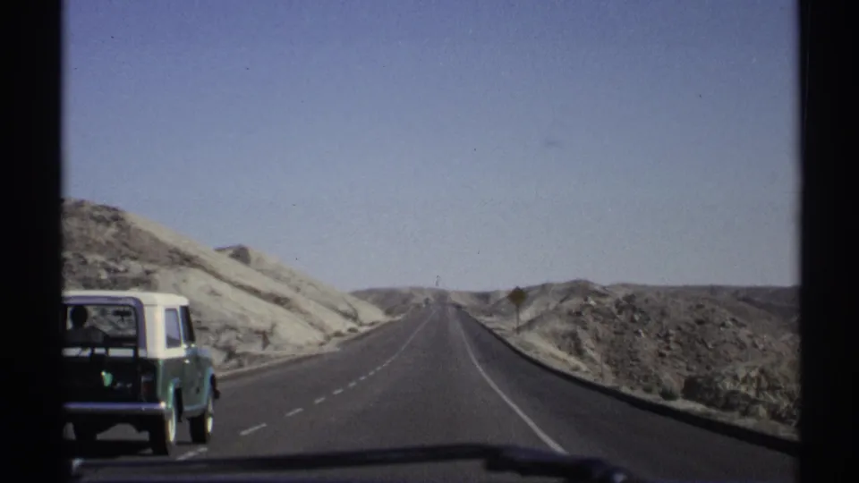 1972 Arizona home-movie frame showing a green and white station wagon on a desert highway between pale hills.