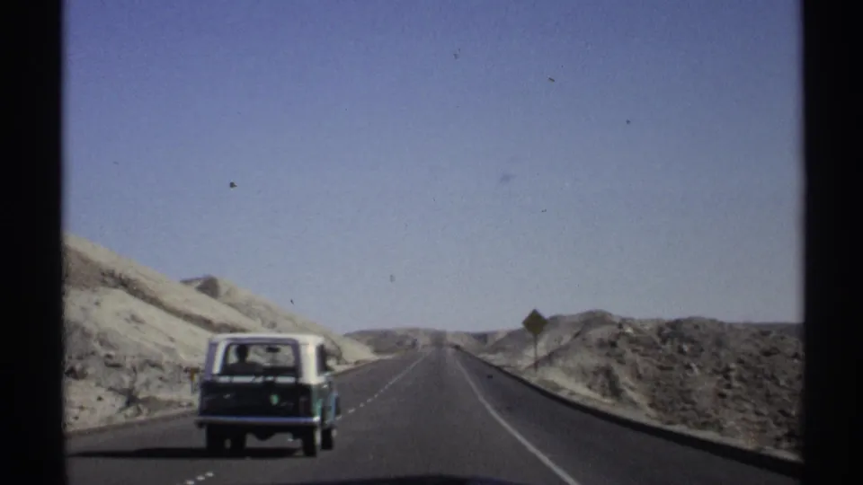 1972 Arizona road-trip footage with a station wagon receding along a desert highway near a roadside warning sign.