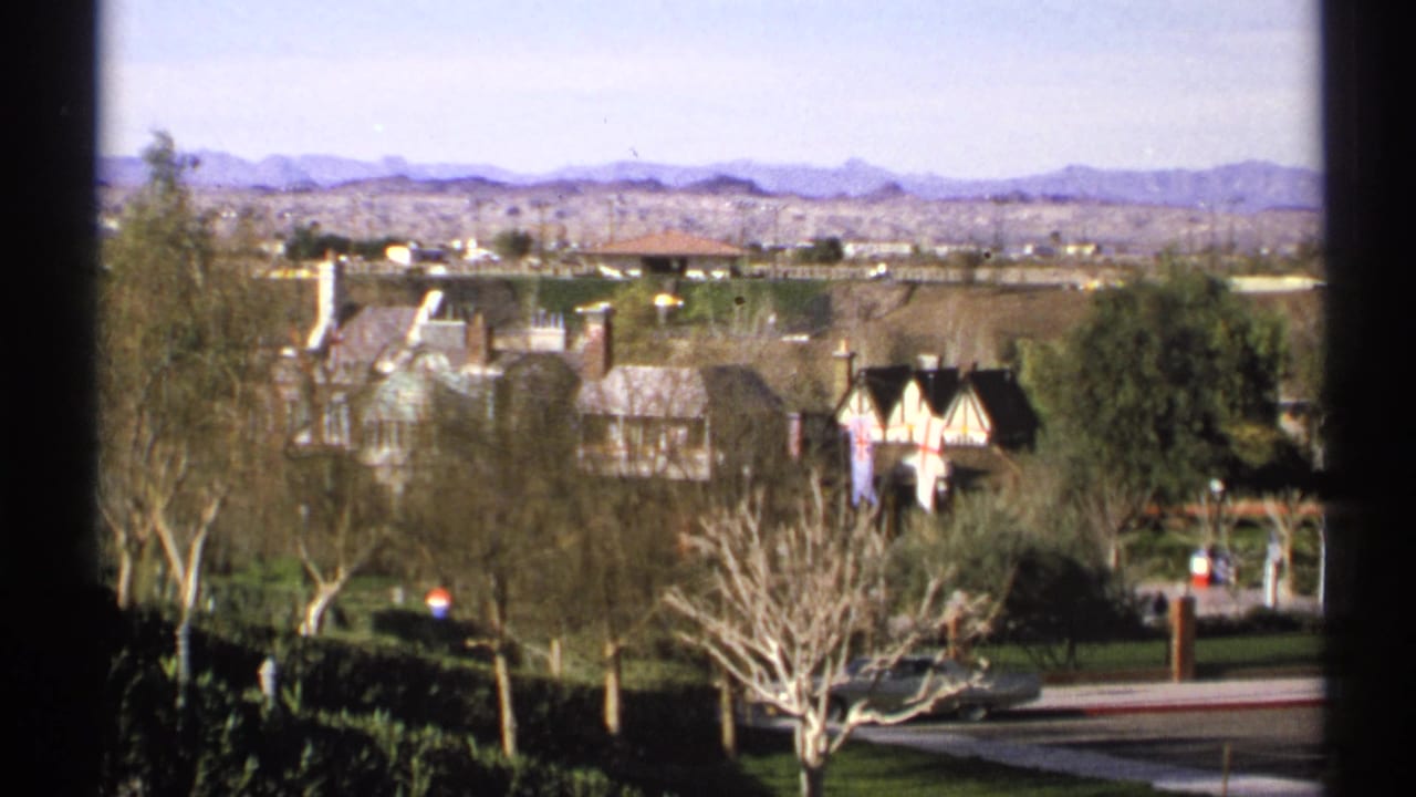 1972 Arizona 8mm view over English-style village buildings with green lawns, bare trees, roadway, and mountains beyond.
