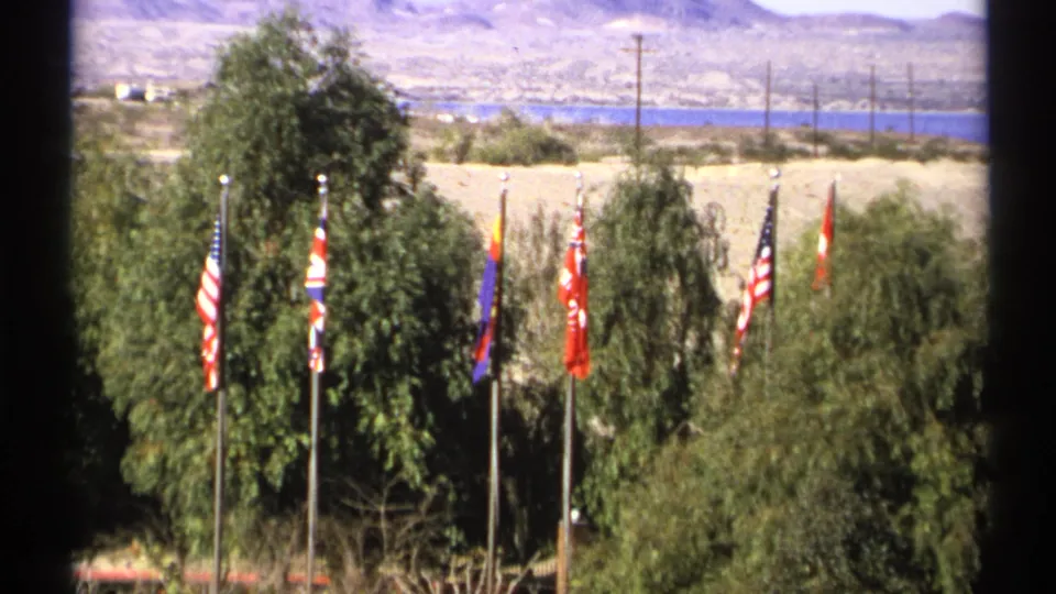 1972 Arizona 8mm frame showing national flags, tall trees, desert terrain, utility poles, and a blue lake or river beyond.
