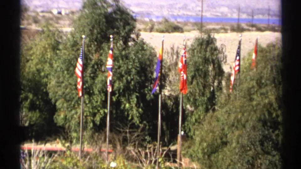 1972 Arizona home-movie view of a flag row framed by trees, with blue water and desert mountains in the background.