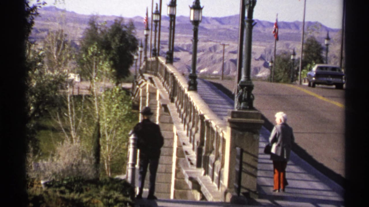 Two adults stand near a stone bridge walkway with lampposts, road, car, trees, and desert mountains.