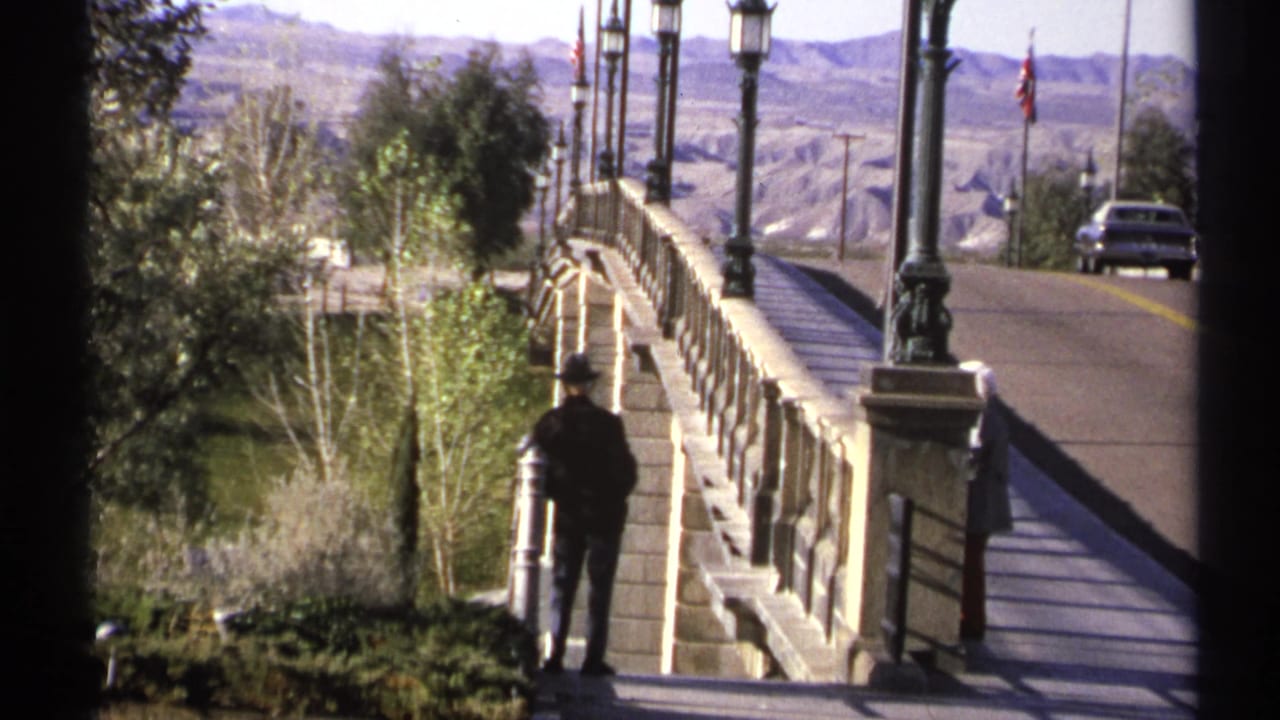 1972 Arizona home-movie frame with a dark-clothed adult beside a bridge railing, street lanterns, road, car, trees, and arid hills.