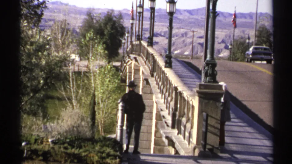 1972 Arizona home-movie frame with a dark-clothed adult beside a bridge railing, street lanterns, road, car, trees, and arid hills.