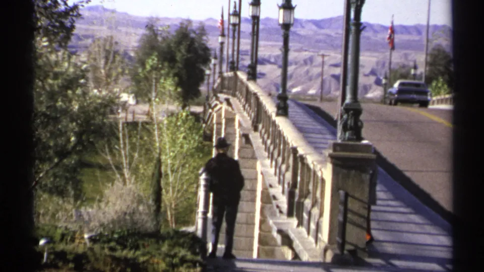 1972 Arizona frame showing a lone adult near the stone bridge rail, with trees, ornate lampposts, road, and distant desert terrain.