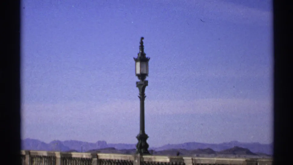 1972 Arizona 8mm frame centered on an ornate street lantern above a bridge rail, with clear sky and desert mountains behind.