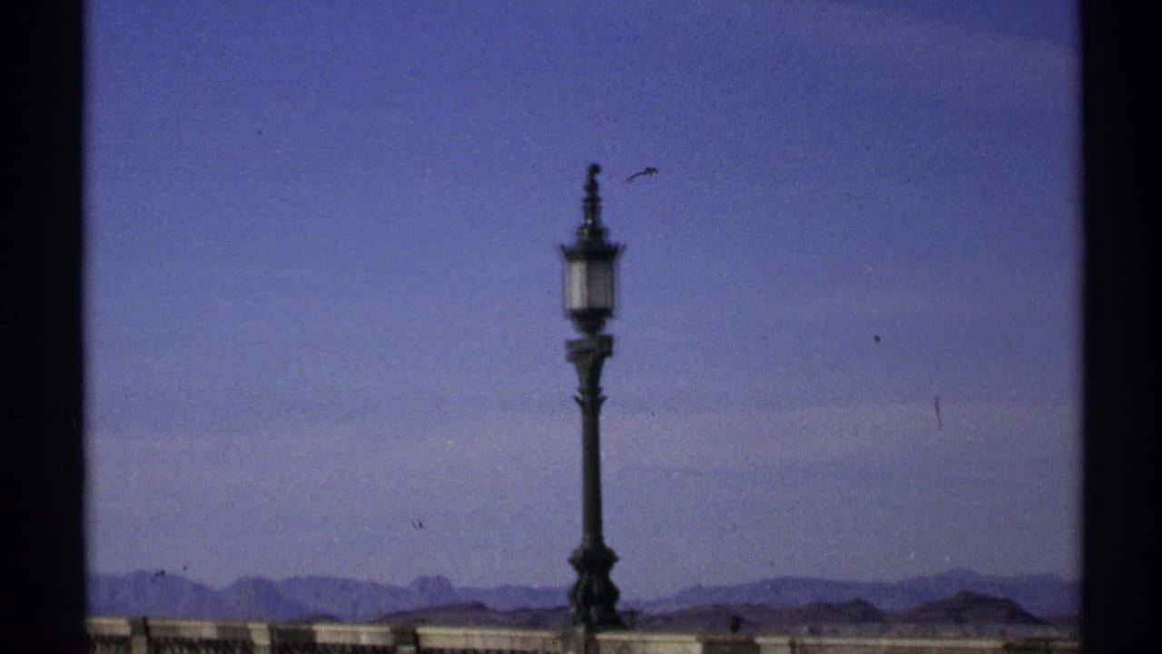Street lamp centered against blue sky with a stone railing and distant mountains.