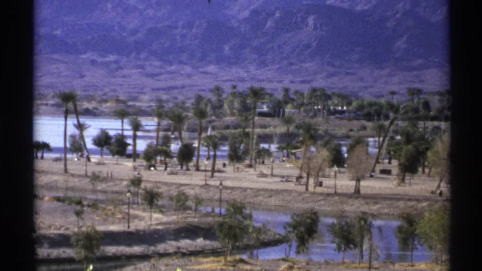 Wide 1972 Arizona 8mm view of a desert oasis area with palms, calm water, dry terrain, and mountains in the background.