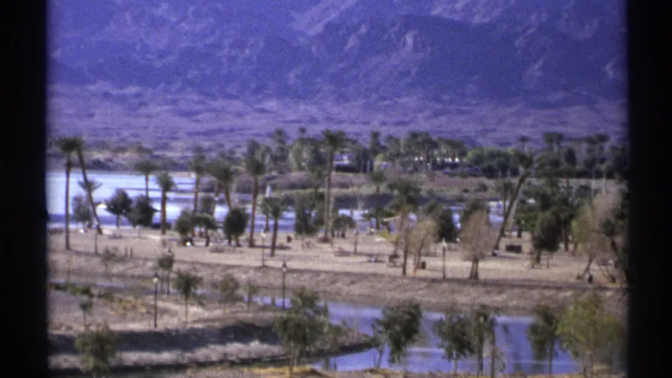 Vintage 8mm frame of a Lake Havasu area landscape, with water, palms, dry terrain, and mountains beyond.