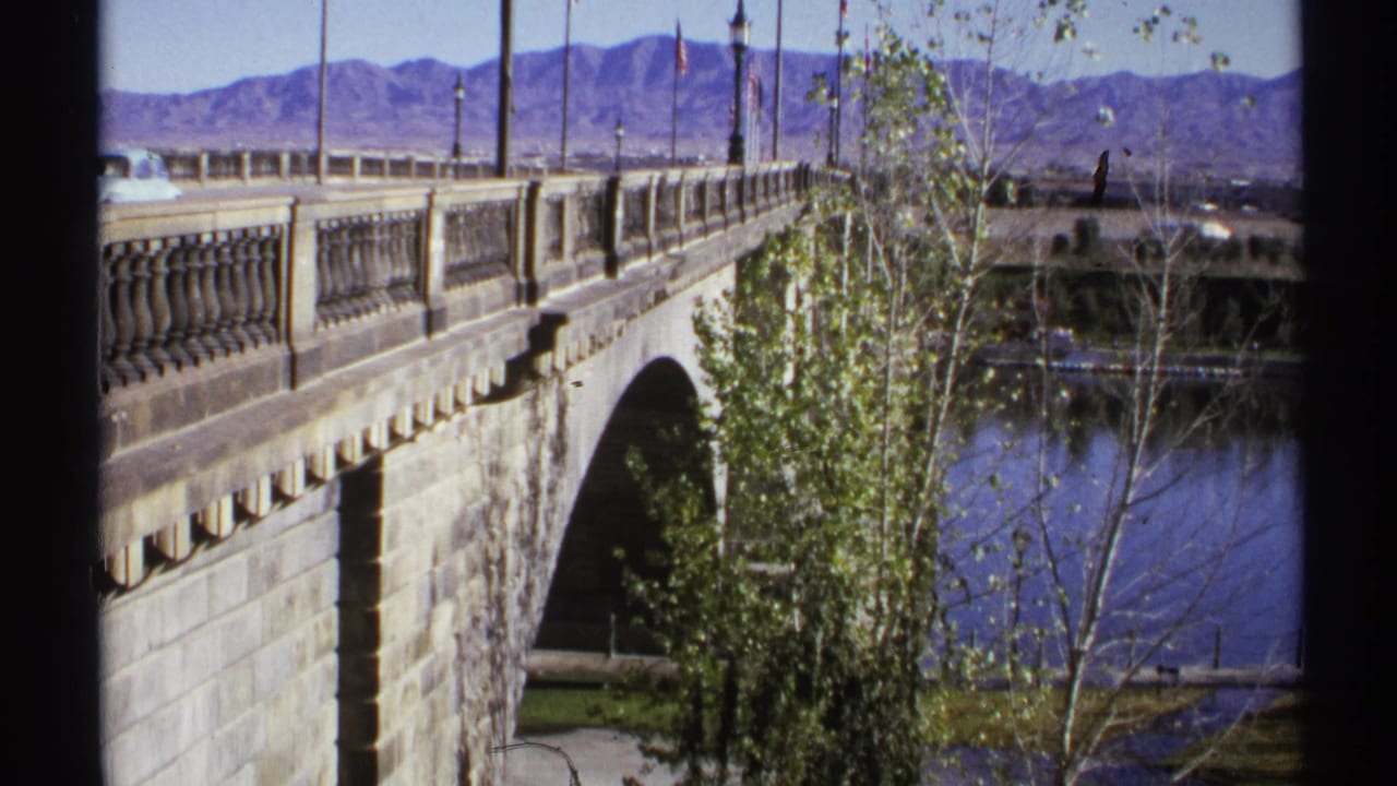 Stone bridge with balustrade over a river, trees in front, and mountains beyond.