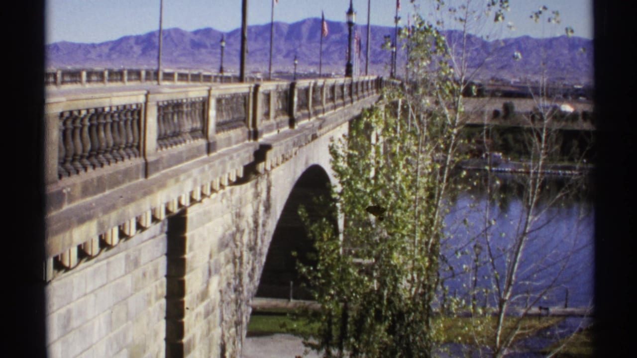 Bridge railing and arch above calm water, with trees and mountains in the background.