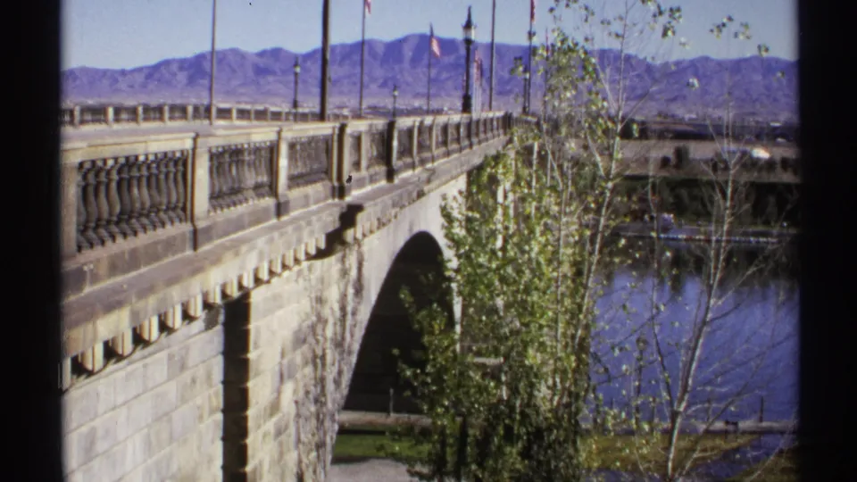 1972 Arizona 8mm view looking along the stone bridge toward flags and mountains, with river water and trees below.