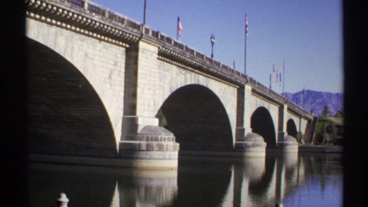 Stone arch bridge over calm water with flags and mountains beyond.