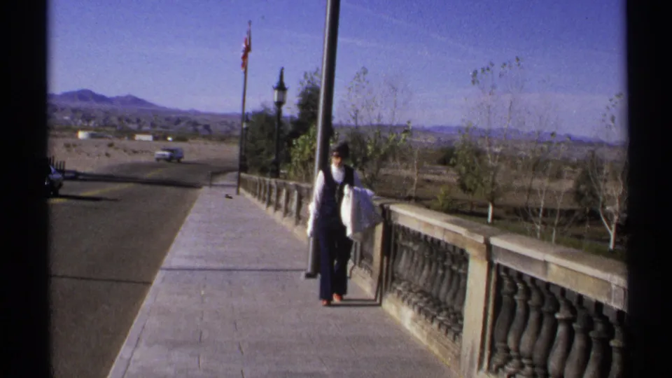 1972 Arizona frame showing an adult man on the bridge walkway, carrying a white bag beside a stone balustrade with mountains in the background.
