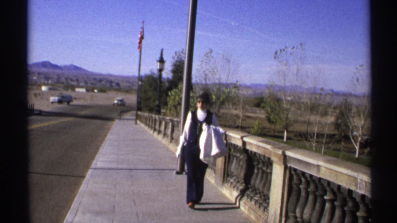 The woman continues along the Arizona bridge walkway, carrying a white bag as cars pass on the road below the mountains.