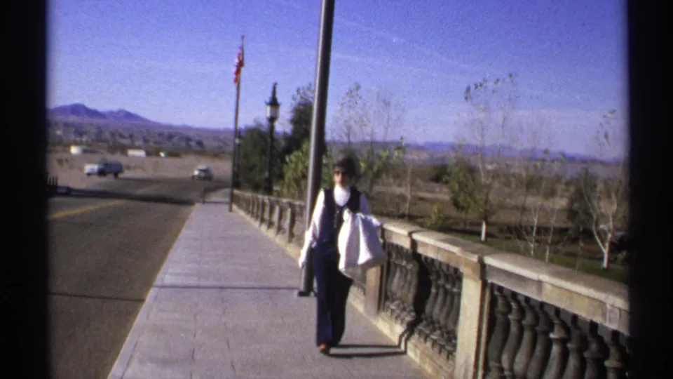 The woman continues along the Arizona bridge walkway, carrying a white bag as cars pass on the road below the mountains.