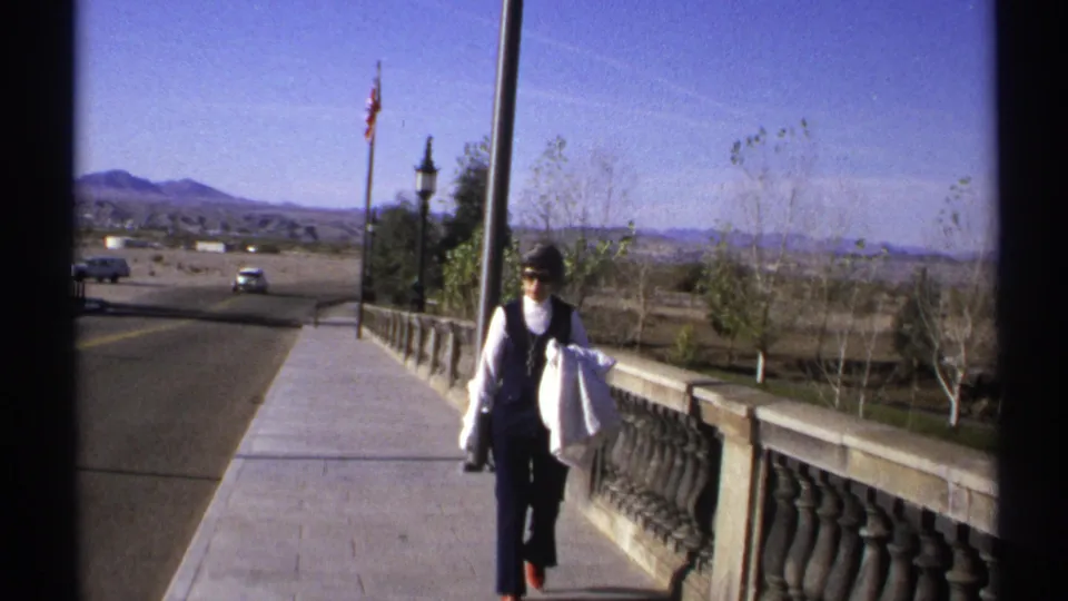 A woman in dark pants and vest walks toward the camera on the Arizona walkway, with a car, balustrade, trees, and mountains visible.