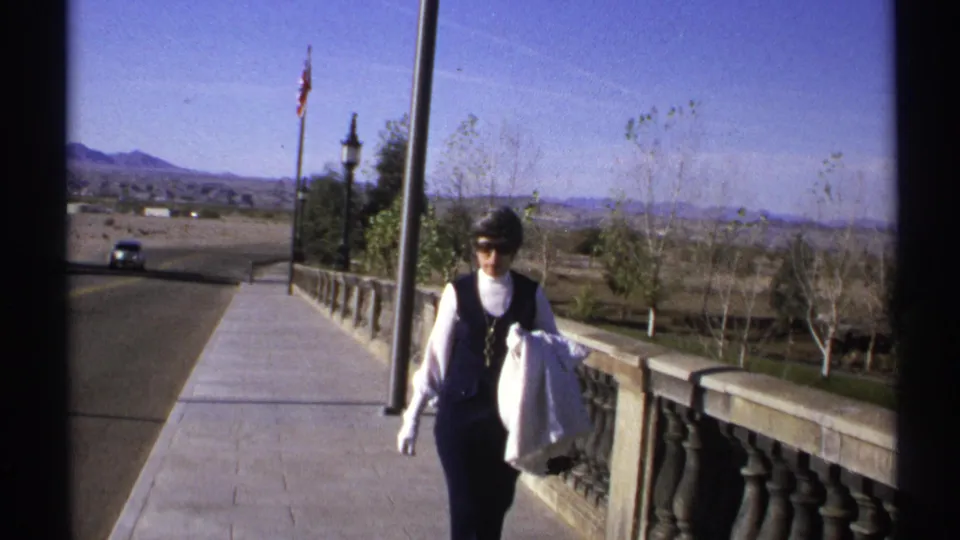 Adult woman in 1970s clothing walks along a stone balustrade in Arizona with road, flagpole, trees, and desert mountains behind her.