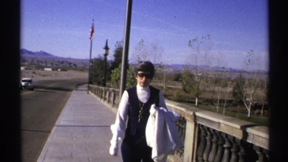 Adult woman wearing sunglasses and gloves walks toward the camera on an Arizona bridge walkway with a road and desert hills behind her.