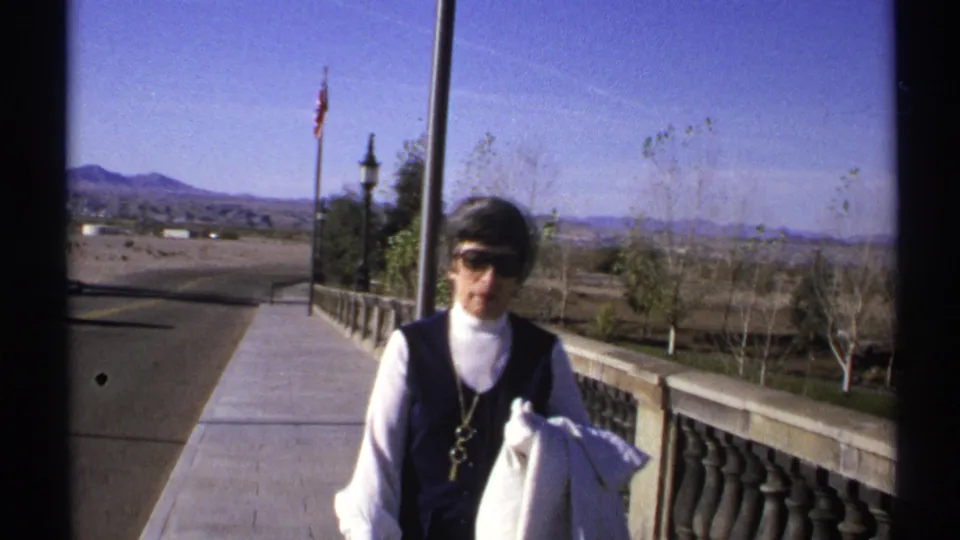 In this 1972 Arizona frame, a woman in a dark vest and white turtleneck walks beside a stone railing with roadway and mountains visible.