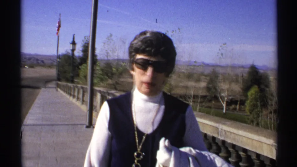 Close 8mm frame of the adult woman in sunglasses, white turtleneck, dark vest, and gloves on the Arizona walkway.