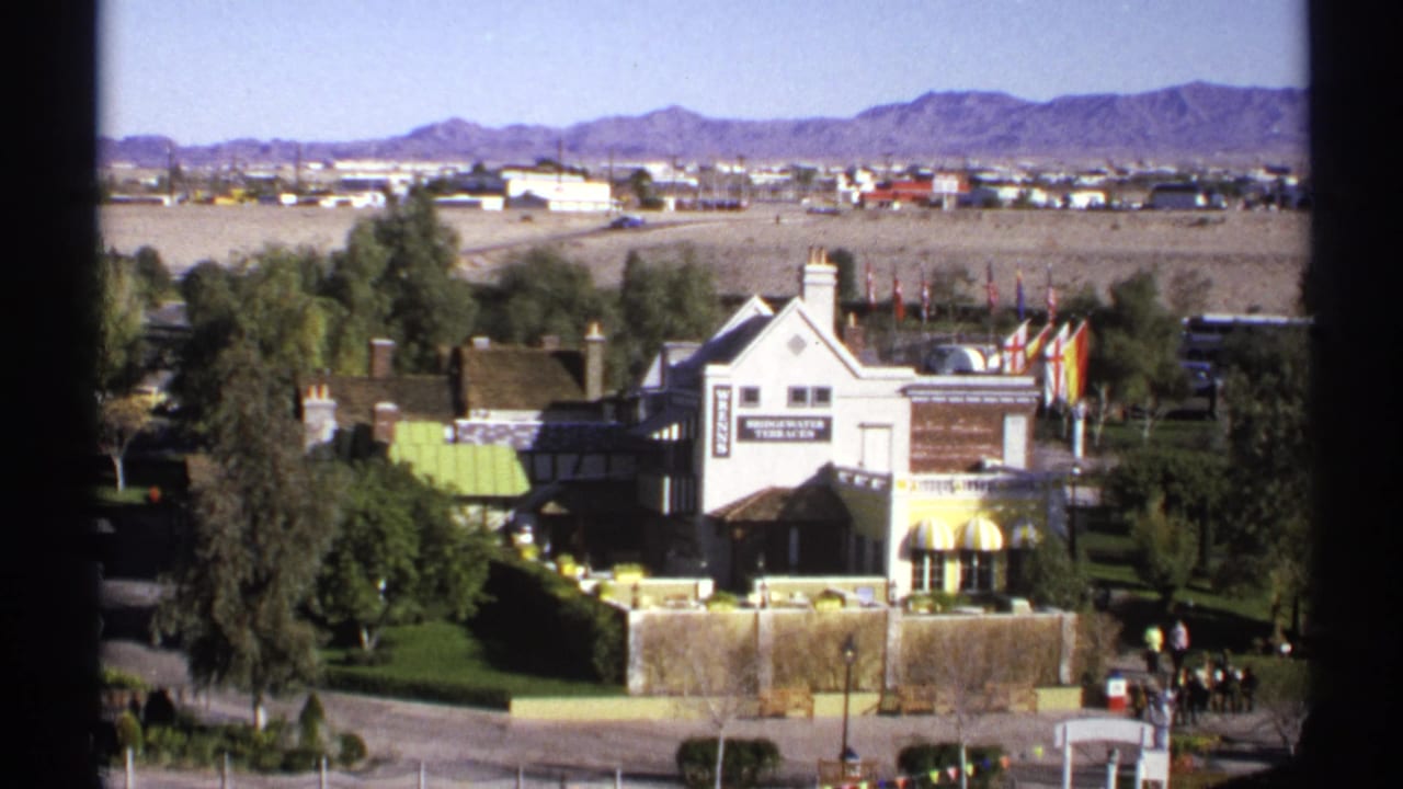 High-angle view of a white Tudor-style building with trees, lawns, signage, and desert mountains beyond.