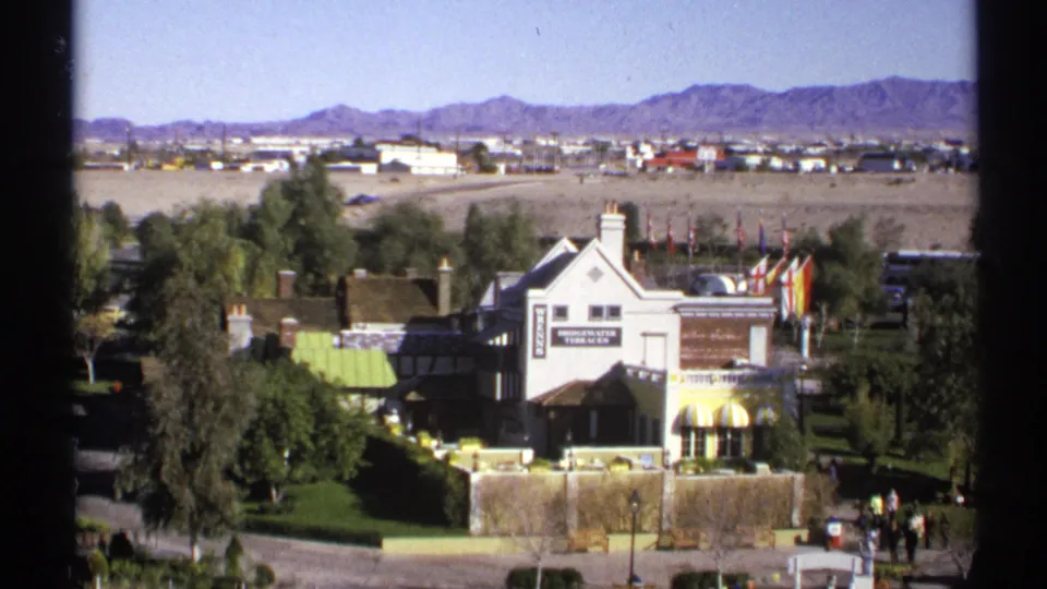 1972 Arizona 8mm view looking down toward Bridgewater Terraces, with manicured greenery, signage, distant development, desert flats, and mountains.