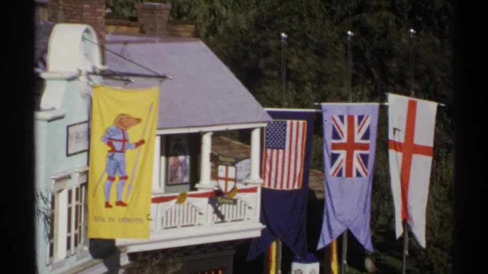 1972 Arizona 8mm view of a flag-decorated miniature village building, with a cartoon dog banner at left and trees behind.